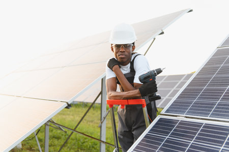 African american technician wearing a hard hat, holding a drill, and installing solar panels on a sunny dayの写真素材