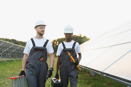 Two diverse engineers carrying tools and a ladder, working on a large solar panel array at a...