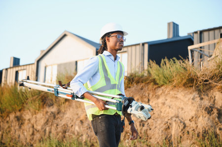 African american engineer or land surveyor working outdoors, carrying a total station and tripod for site developmentの写真素材