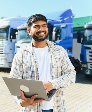 Indian man smiling, holding a laptop, standing in front of a truck fleet. He is managing logistics...