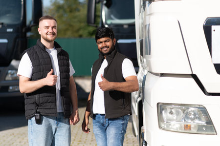Two smiling men, European and Indian, standing together at a truck parking, showing thumbs up,...