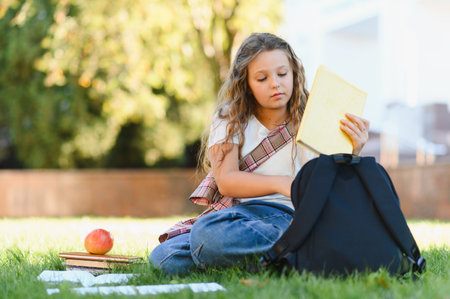 Student girl sitting on grass holding a book near her backpack, enjoying quiet reading time outside...