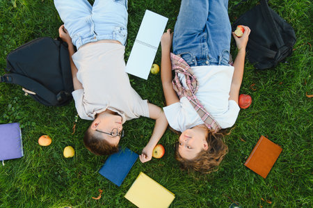 Two young students lying on green grass outdoors, having a break with books and a backpack while holding handsの写真素材