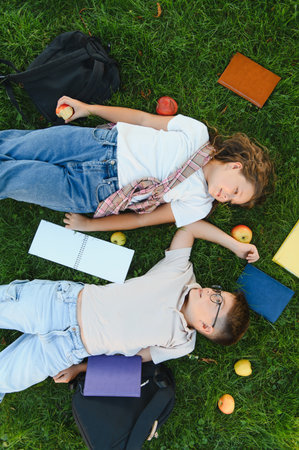 Boy and girl students lying on green grass, enjoying a break with books, apples, and backpacks. Top view of friendshipの写真素材