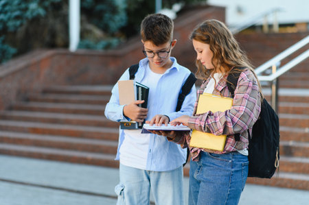 Schoolchildren boy and girl with backpacks and books discussing studies outdoors near school building, learning and teamworkの写真素材