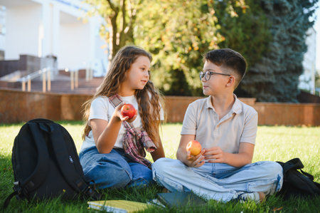 Happy schoolchildren sitting on green grass eating fresh apples. Friends conversing during school break outdoors, promoting healthy lifestyleの写真素材