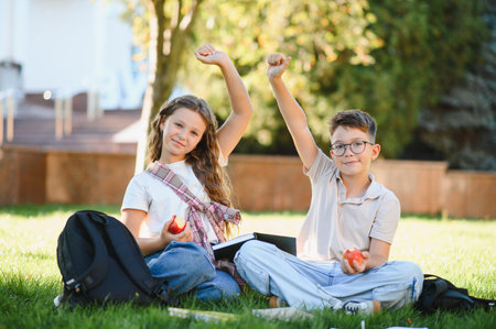 Happy schoolchildren sitting on green grass, raising hands, having fun, studying and celebrating success during school timeの写真素材