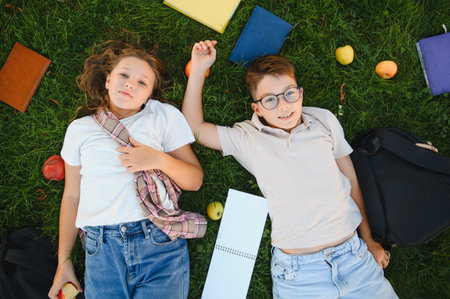 Two happy schoolchildren lying on green grass with books, backpacks, and apples around them,...
