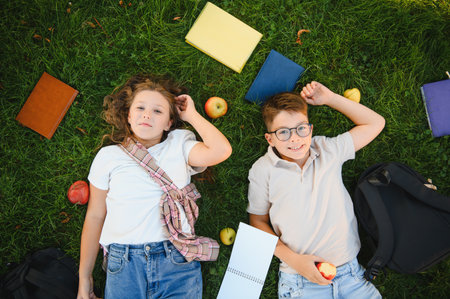 Schoolchildren lying on green grass enjoying free time, surrounded by school supplies and apples,...