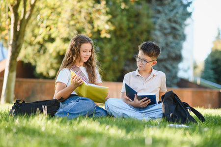 Two young students sitting on the grass, holding books, and studying outdoors. Learning and...