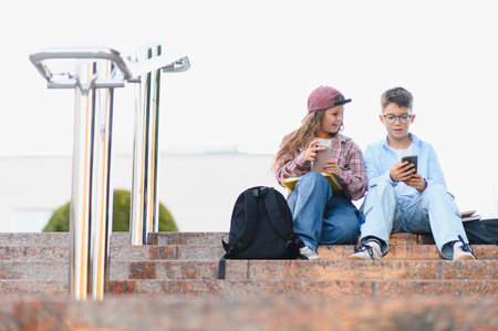 Two schoolchildren sitting on campus stairs, boy showing something on smartphone to girl. Building...