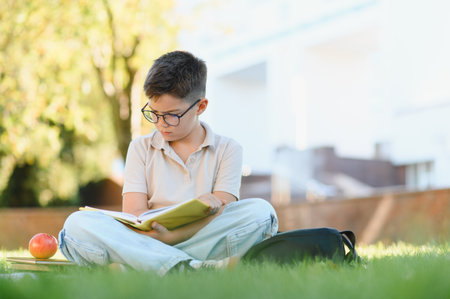 Young schoolboy with glasses attentively reading an open book while sitting cross legged on green...