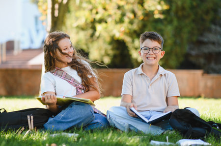Two cheerful schoolchildren sitting on green grass, reading books and smiling. Learning outdoors,...
