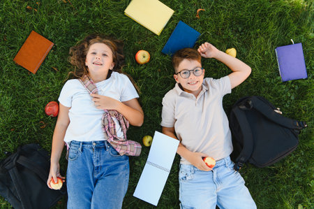 Schoolchildren lying on green grass, smiling, holding apples, with backpacks and books around them,...