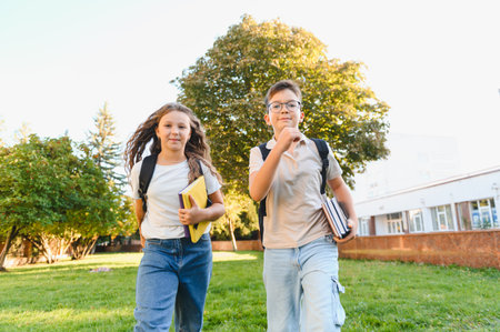 Two cheerful schoolchildren with backpacks and books running across a grassy campus lawn, heading to...