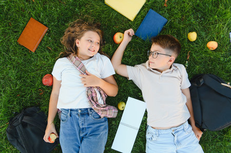 Two cheerful schoolchildren lying on green grass with books and backpacks, enjoying a break from...