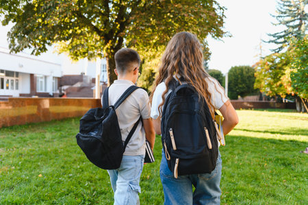 Two schoolchildren, a boy and a girl, walking away from the camera on a grassy school campus towards a buildingの写真素材