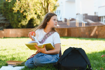 Happy schoolgirl reading book and eating apple while sitting on green grass. Backpack and textbooks...