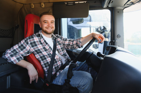 Smiling young truck driver in plaid shirt and seatbelt at steering wheel of semi truck cab,...