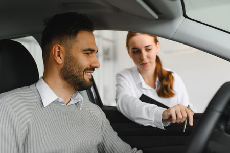 Saleswoman assisting a smiling male customer sitting in a car, discussing vehicle features at a...
