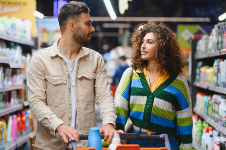 Young couple walking through the supermarket aisle pushing a shopping cart, choosing products for...