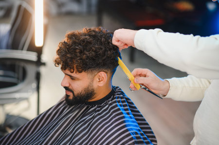 Barber styling curly hair of male client in modern barbershop, professional grooming and precise...