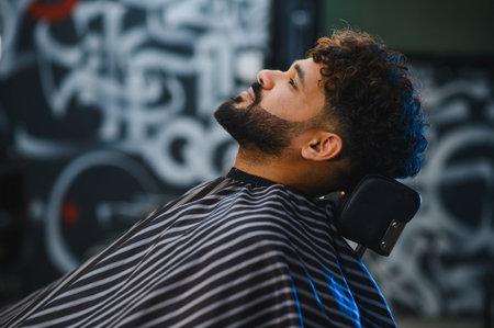 Man relaxing in a barbershop chair with a striped cape, enjoying a professional grooming service for...