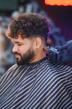 Man with curly hair getting a fresh fade haircut from a barber wearing gloves, focusing on stylish...
