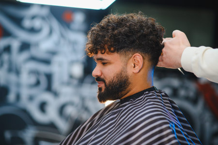 Man with curly hair receiving a stylish fade haircut and beard trimming service from a professional...