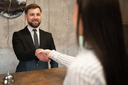 Friendly hotel receptionist in a suit smiling, shaking hands with a arriving guest at the reception desk, offering hospitalityの写真素材