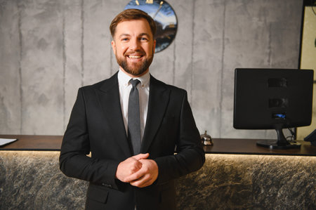 Friendly hotel receptionist with a beard smiling, standing at a modern reception desk in a lobby, providing hospitality serviceの写真素材