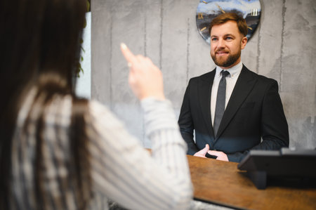 Friendly male receptionist wearing suit and tie assisting a guest at a hotel front desk, providing check in serviceの写真素材