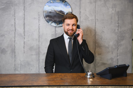 Young man working as a hotel concierge, standing at reception desk, holding phone, offering professional hospitality serviceの写真素材