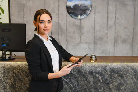 Professional woman smiling, holding paperwork while providing excellent hospitality service at a hotel reception counterの写真素材