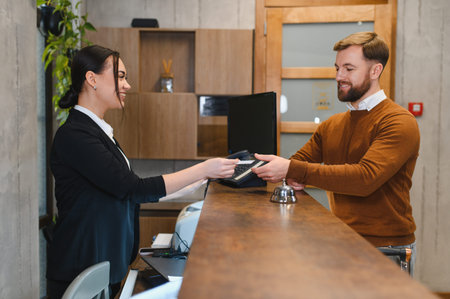 Administrator giving key card to smiling male guest at a hotel reception, providing hospitality service during check inの写真素材
