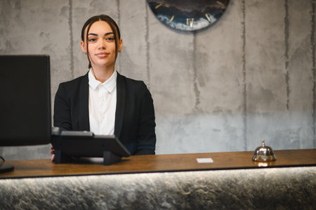 Young woman smiling, standing behind hotel reception desk. Providing hospitality service in a professional workspaceの写真素材