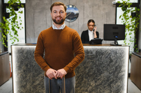Smiling man holding luggage, standing at hotel reception desk, ready for check in. Female receptionist working in backgroundの写真素材