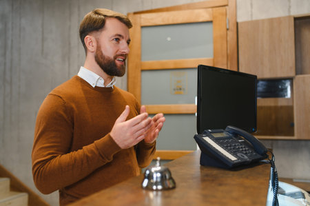 Bearded man working at reception desk, speaking to client. Demonstrating hospitality, service, and professional welcomeの写真素材