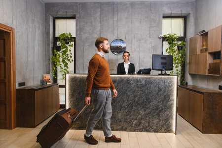 Man with suitcase arriving at a modern hotel lobby, female receptionist smiling and waiting to welcome guestの写真素材