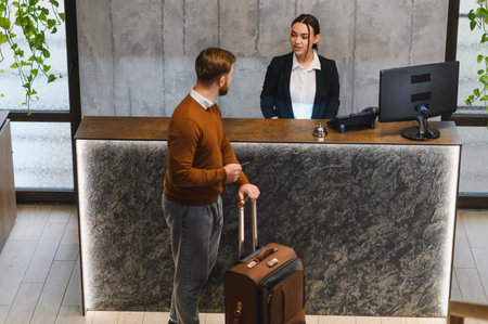 Man with suitcase checking in at modern hotel reception, talking to friendly female administrator. Hospitality serviceの写真素材