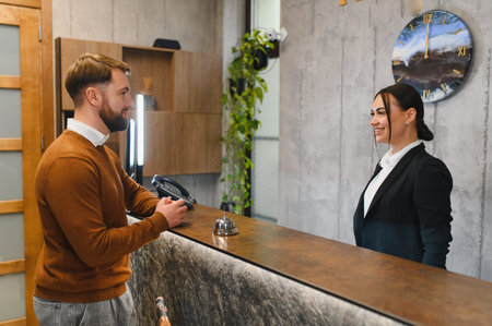 Male customer communicating with smiling female receptionist at modern hotel lobby reception desk, showing hospitality and serviceの写真素材