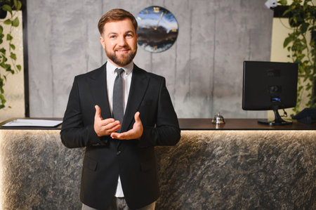 Smiling man in a suit standing at a modern hotel reception desk, presenting a welcoming gesture toward guestsの写真素材