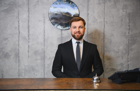 Professional male receptionist standing behind a wooden counter, offering hospitality and service in a hotel lobby settingの写真素材
