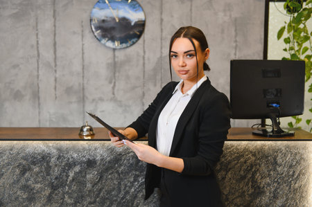 Professional woman standing at a modern reception desk in a hotel lobby, holding a clipboard and looking at the cameraの写真素材
