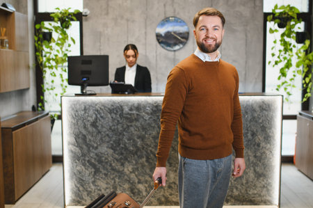 Smiling man pulling luggage, standing in modern hotel lobby, with a female receptionist working at the counterの写真素材