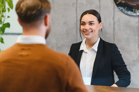 Friendly female receptionist smiling, providing service to a male client at a hotel reception desk, offering hospitality and supportの写真素材