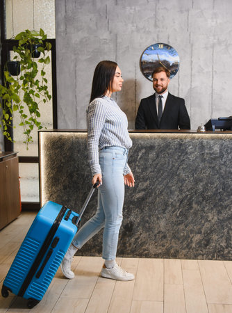 Guest arriving at modern hotel lobby, pulling luggage for check in with a male receptionist standing behind counterの写真素材