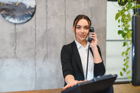 Young female receptionist smiling while answering a landline phone at a modern office front desk, professional and confident serviceの写真素材