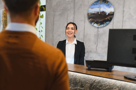 Friendly female receptionist smiling while assisting a male client checking in at a modern hotel lobby reception counterの写真素材