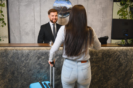Woman with luggage checking into hotel, smiling male receptionist providing reception service and hospitality at the counterの写真素材